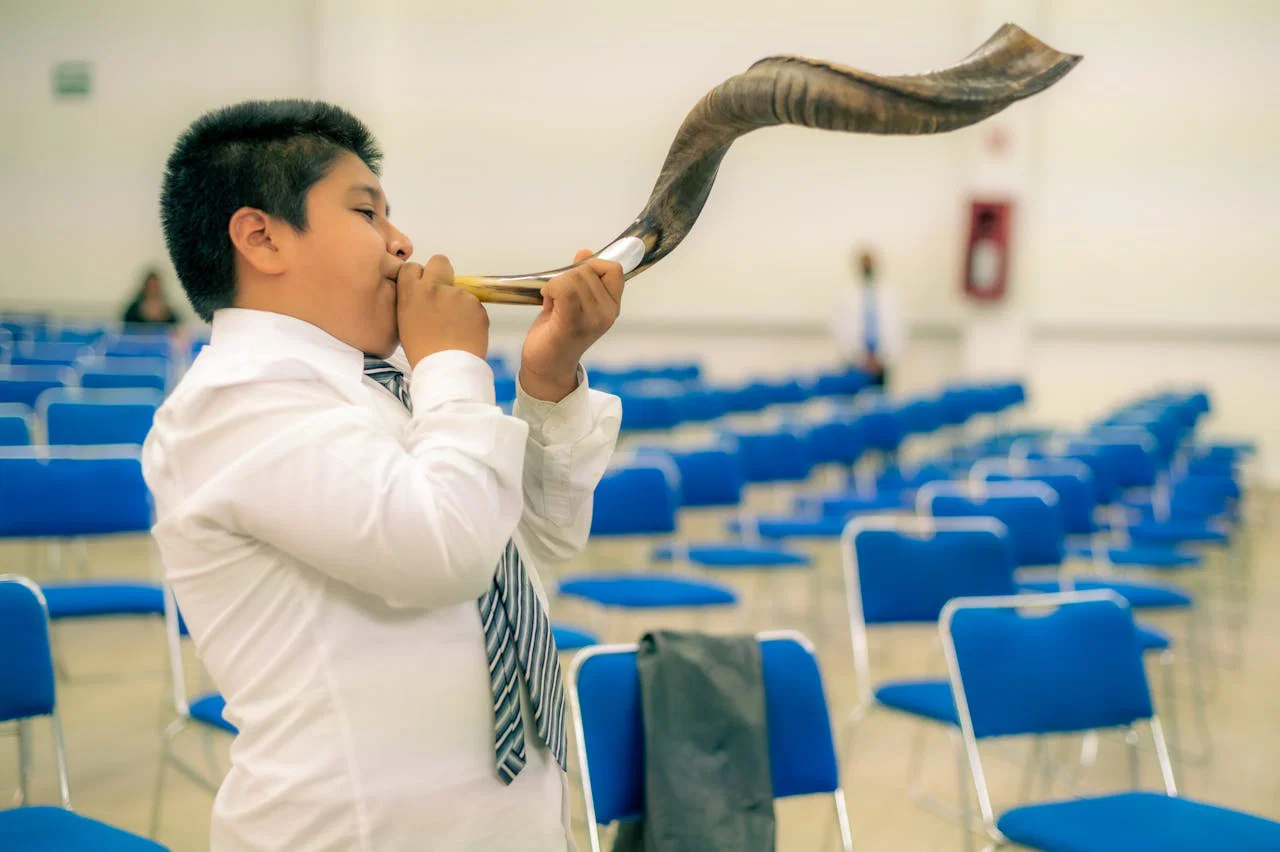 Young boy blowing a shofar in an empty auditorium, dressed in formal attire.